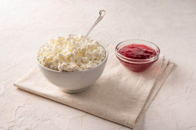 Cottage cheese in a bowl with a spoon and fruit jam on a linen napkin on a light background.