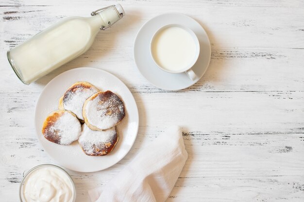 Cottage cheese pancakes. Cheesecakes. Syrniki or sirniki. Healthy breakfast. White dishes. Bottle of milk. A cup of milk. Yogurt or sour cream. White background. Top view. Space for text