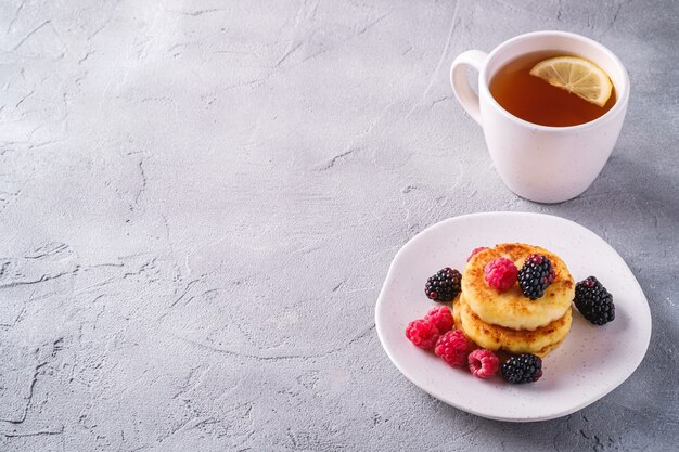 Cottage cheese pancakes, curd fritters dessert with raspberry and blackberry berries in plate near to hot tea cup with lemon slice on stone concrete background, angle view copy space