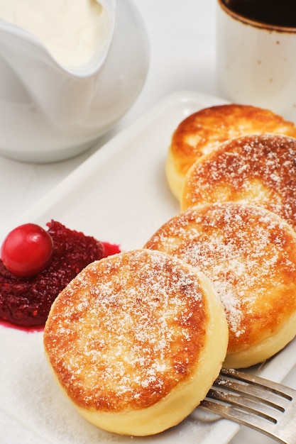 Cottage cheese pancakes or fried cottage cheese pancakes, sprinkled with powdered sugar, on a plate with cranberry jam and sour cream.