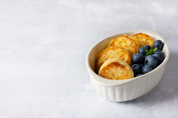 Cottage cheese pancakes on a light background. Syrniki with fresh blueberries. Pancakes with cottage cheese on a white dish. Copy space for your text.