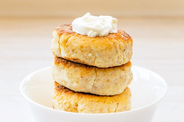 Photo cottage cheese pancakes stack with sour cream in round white bowl on white wooden table close up