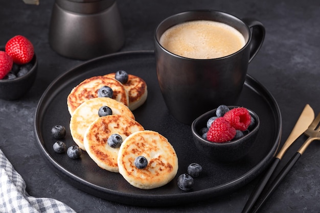 Photo cottage cheese pancakes with fresh with fresh raspberries and blueberries on dark ceramic plate cup ...