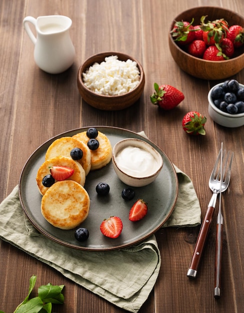 Cottage cheese pancakes with sour cream milk blueberries and strawberries on a dark wooden background Healthy and delicious breakfast Rustic style