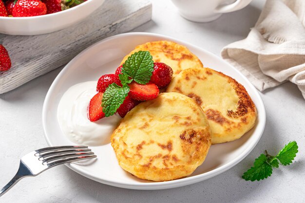 Cottage cheese pancakes with strawberries and cream in a white plate on a light gray background closeup