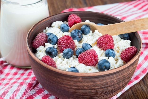 Cottage cheese with raspberries and blueberries in brown clay bowl and wooden spoon on red checkered towel