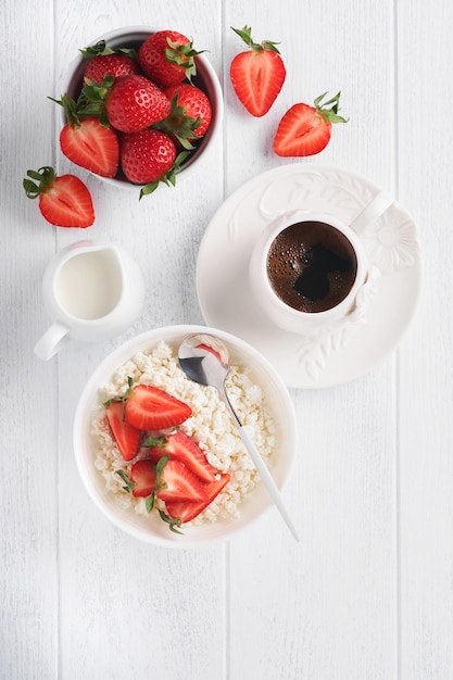 Cottage cheese with strawberries Breakfast from cottage cheese with slices fresh strawberries cream cup of coffee in white bowl on white wooden background Top view Food concept Mock up