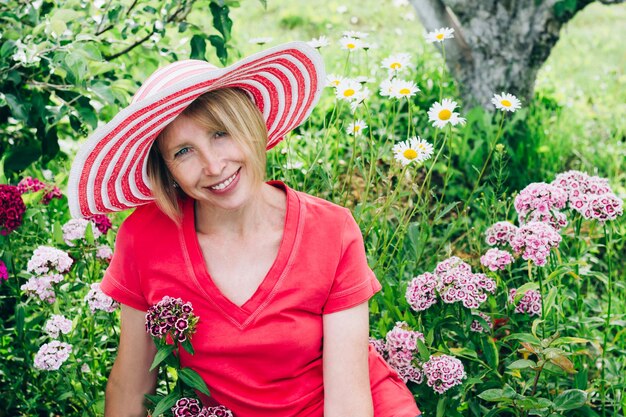 Cottagecore en Staycation thuis. Gelukkig oudere blonde vrouw in hoed rusten tussen bloemen. Ontspan in isolement.