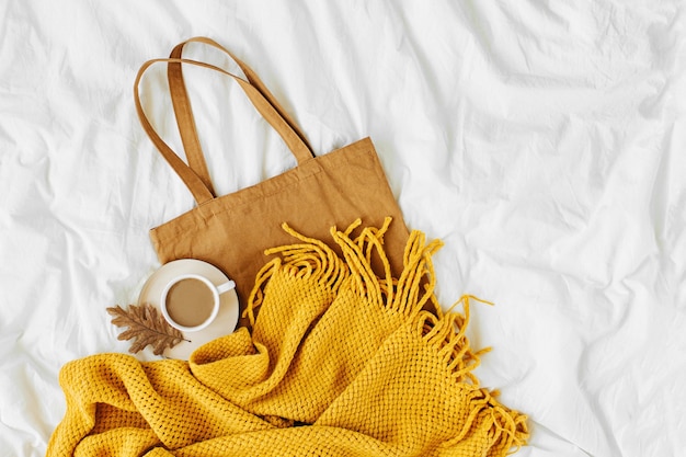 Cotton eco bag with cup of coffee and yellow knitted scarf on\
bed. autumn concept.