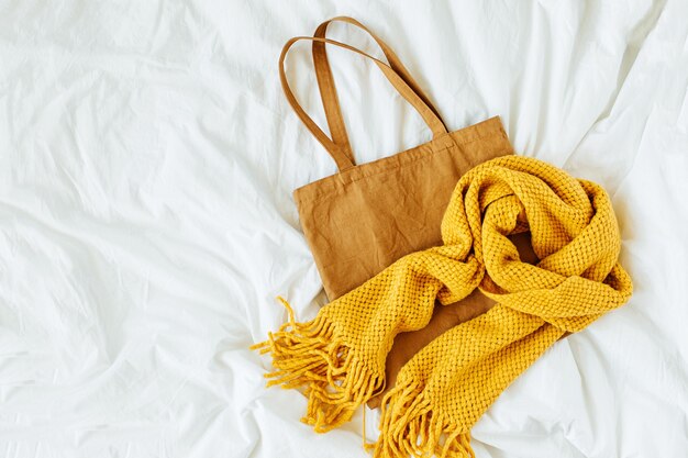 Cotton eco bag with warm yellow knitted  scarf on bed. Autumn fashion concept.