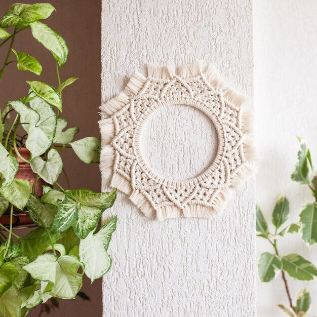 緑の葉と白い壁に掛かっている綿のマクラメ曼荼羅の壁の装飾手作りのマクラメの花輪天然綿糸エコ家の装飾