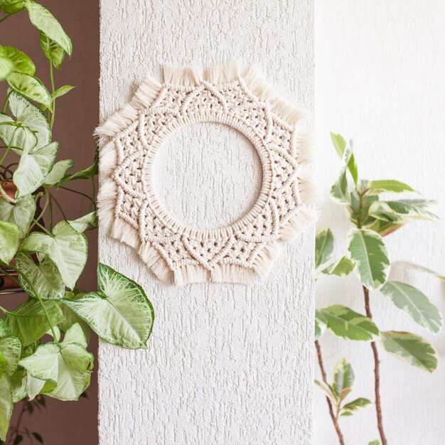 Cotton macrame mandala wall decoration hanging on white wall with green leaves Handmade macrame wreath Natural cotton thread Eco home decor