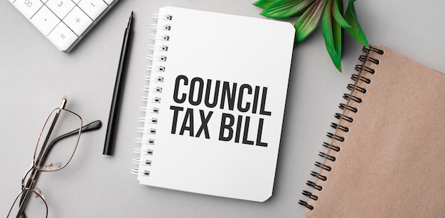 CoUNCIL TAX BILL is written in a white notebook with calculator, craft colored notepad, plant, black marker and glasses