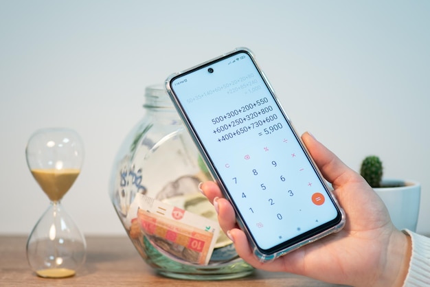 Photo counting saved money for a purchase phone with calculator in female hands with jar with saved up money on the background