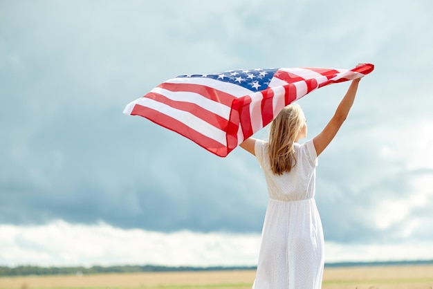 country, patriotism, independence day and people concept - happy young woman in white dress with national american flag on cereal field