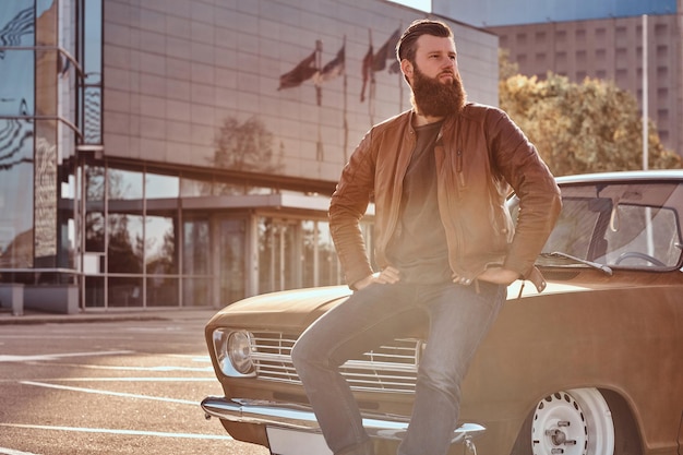 Country style concept. Stylish male leaning on tuned retro car in the city parking near skyscraper at bright sunset.