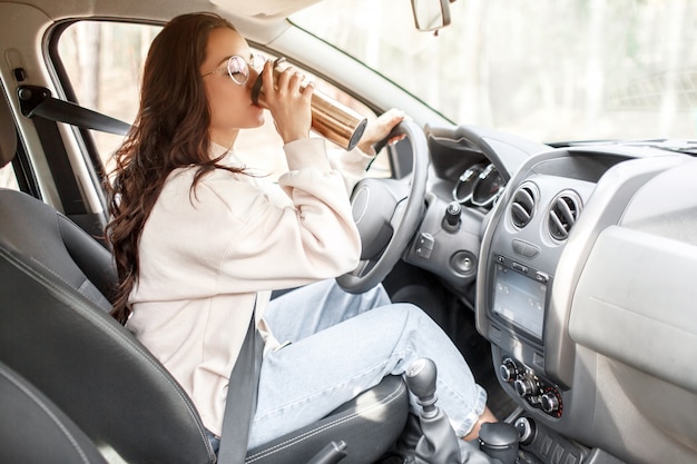 Country trip traveling by car. Female model drinks coffee or tea from a thermos.