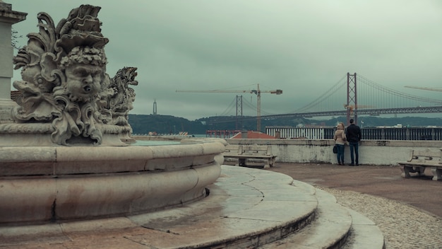 La coppia ammira il ponte il 25 aprile e la statua di cristo salvatore dal ponte di osservazione. vecchia fontana con fantasiose sculture di pesce sul punto di vista largo das ecessidades lisbona portogallo
