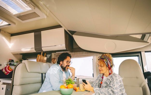 Couple of adult tourist enjoy leisure time together inside a\
modern camper van renting motor home for summer travel holiday\
vacation van life style for young people woman using phone and man\
eating