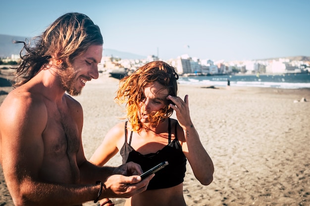 Couple of adults at the beach talkin and loking at the phone of the woman stand up on the sand - woman in bikini looking at ther phone and a man looking at the same phone