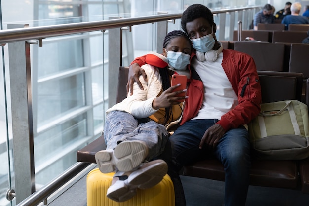 Couple of african tourists wear face masks and sit together on chair in airport waiting for flight