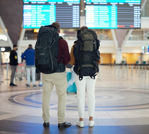 Couple airport and reading on board schedule and screen for flight on travel plane or adventure with back Man woman and global immigration in lobby for airplane time departure or transportation