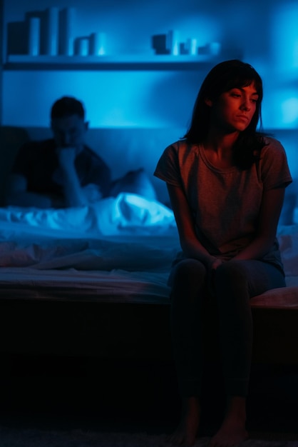 Couple argument Night fight Divorce breakup Dissatisfied sad pensive woman sitting on bed on distance from thoughtful man in dark red blue neon light on defocused background