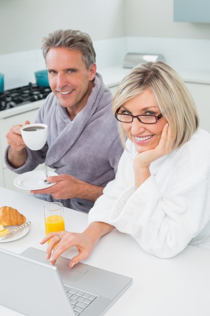 Couple in bathrobes with coffee and juice using laptop in kitchen