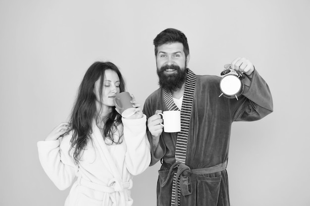 Couple in bathrobes with mugs Man with beard and sleepy woman enjoy morning coffee or tea Time to wake up and have nice day Guy husband hold coffee and clock Every morning begins with coffee