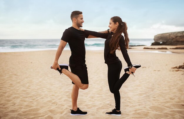 Couple in black sportswear training by seaside doing stretching exercises for legs standing in front of each other