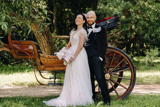 A couple of the bride and groom are standing near the carriage in nature in retro style