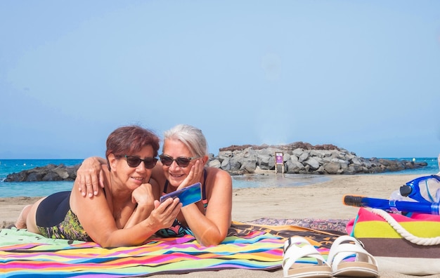 Couple of Caucasian mature women girlfriends lying sunbathing on sea beach using mobile phone elderly females enjoying free time and vacation horizon over water