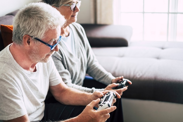 Couple of caucasian retired seniors playing videogames together\
at home sitting on the sofa - mature woman and man married - two\
get engaged forever - holding a controller or a joystick