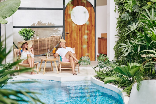 Couple celebrating anniversary in spa hotel, they are sunbathing by swimming pool, drinking cocktails and talking