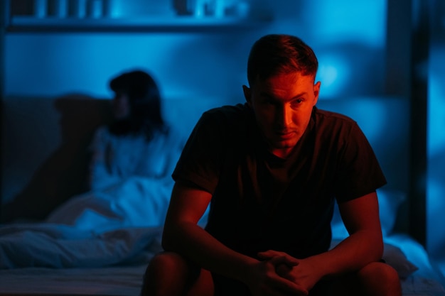 Couple conflict Domestic abuse Bad relationship Aggressive man sitting on bed with afraid insecure woman victim in dark night red blue neon light on defocused background