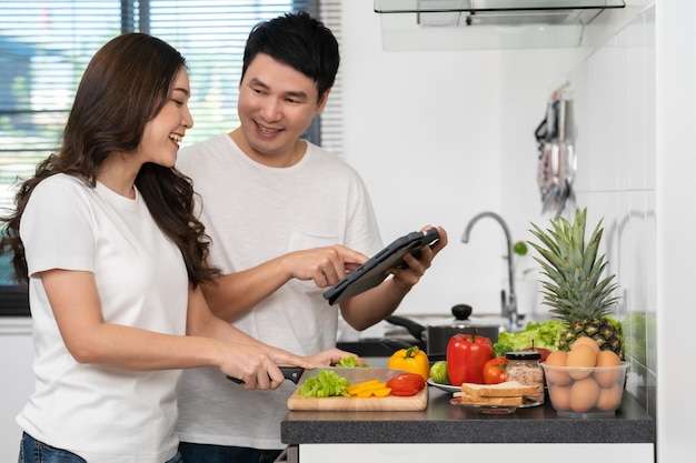 自宅のキッチンにあるタブレットコンピューターでレシピに従って野菜を調理し、準備するカップル