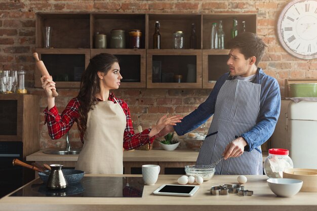 Couple cooking pastry and quarreling in their loft kitchen at home. Man and woman arguing while preparing dough, woman swing with rolling-pin, domestic violence concept, copy space