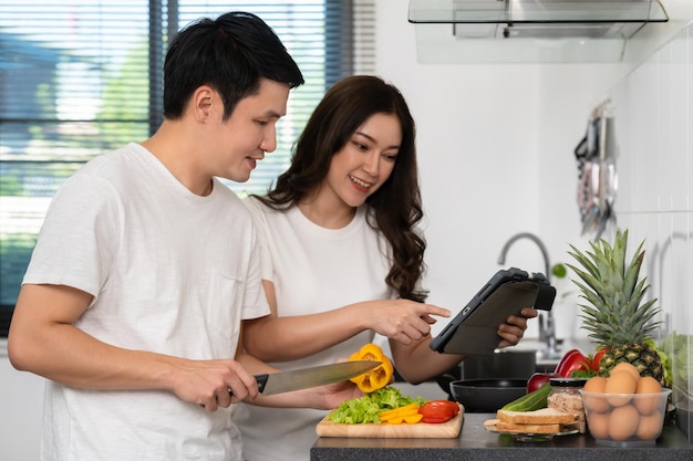 自宅のキッチンにあるタブレットコンピューターでレシピに従って野菜を調理し、準備するカップル