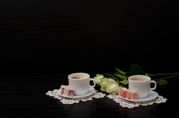 Un paio di tazze di caffè con latte, turkish delight, un bouquet di rose bianche su sfondo nero. spazio per il testo