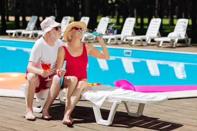 Couple of cute fashionable pensioners blowing soap bubbles while sitting near the pool