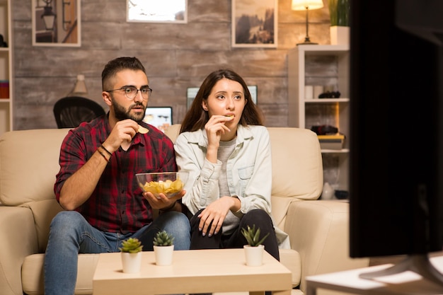 Пара ест чипсы и в шоке смотрит по телевизору во время просмотра фильма в ночное время.