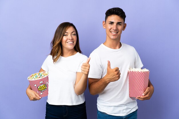 Couple eating popcorn while watching a movie on purple giving a thumbs up gesture with both hands and smiling