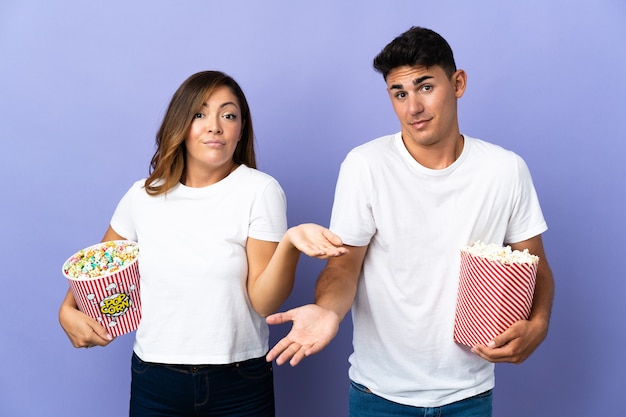 Couple eating popcorn while watching a movie on purple having doubts while raising hands and shoulders