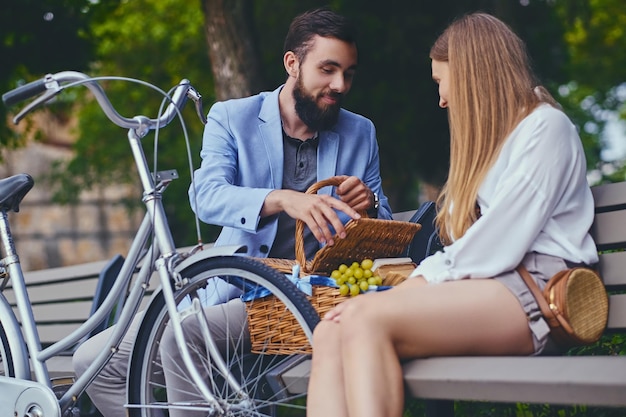 A couple eats grape on a bench in a park.