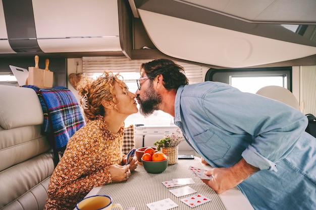 Couple enjoy leisure time playing cards and kissing with love inside a camper van Travel and holiday vacation concept Van life for travel people Young adult man and woman in relationship indoor