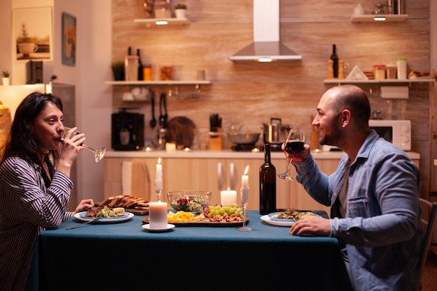 Coppia gustando un bicchiere di vino. coppia felice parlando, seduto al tavolo nella sala da pranzo, godendosi il pasto, celebrando il loro anniversario a casa avendo un momento romantico.