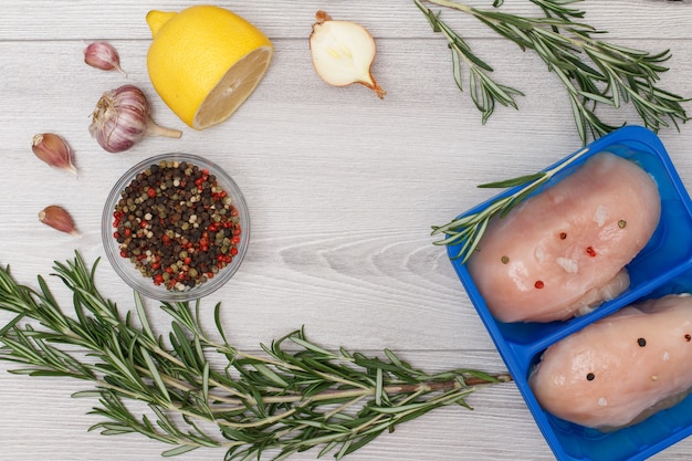 Couple of fresh raw chicken breasts or fillet in plastic container with garlic, onion, lemon, allspice peppers in glass bowl and rosemary on a wooden background. Top view.