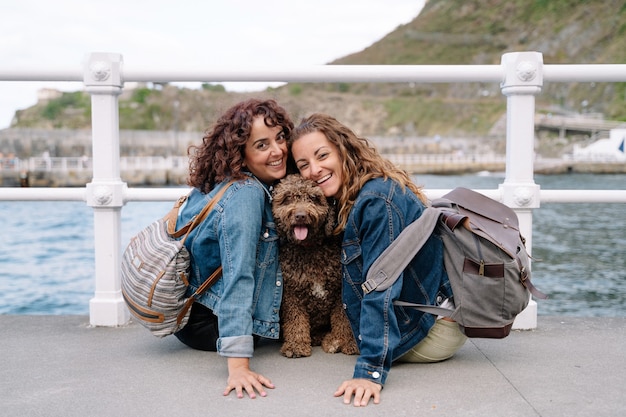 Couple of friends hugging brown water dog. Horizontal view of women traveling with pet. Lifestyle with animals outdoors.