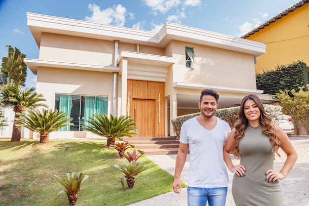 Couple in front of new large modern house, outdoors holding key. Happy couple with dream house in the background.