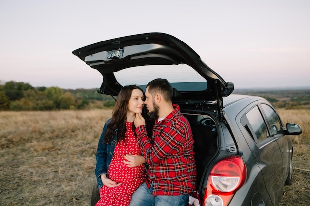 Couple of future young parents sit in the trunk of their family\
car and hug outside the city in red.